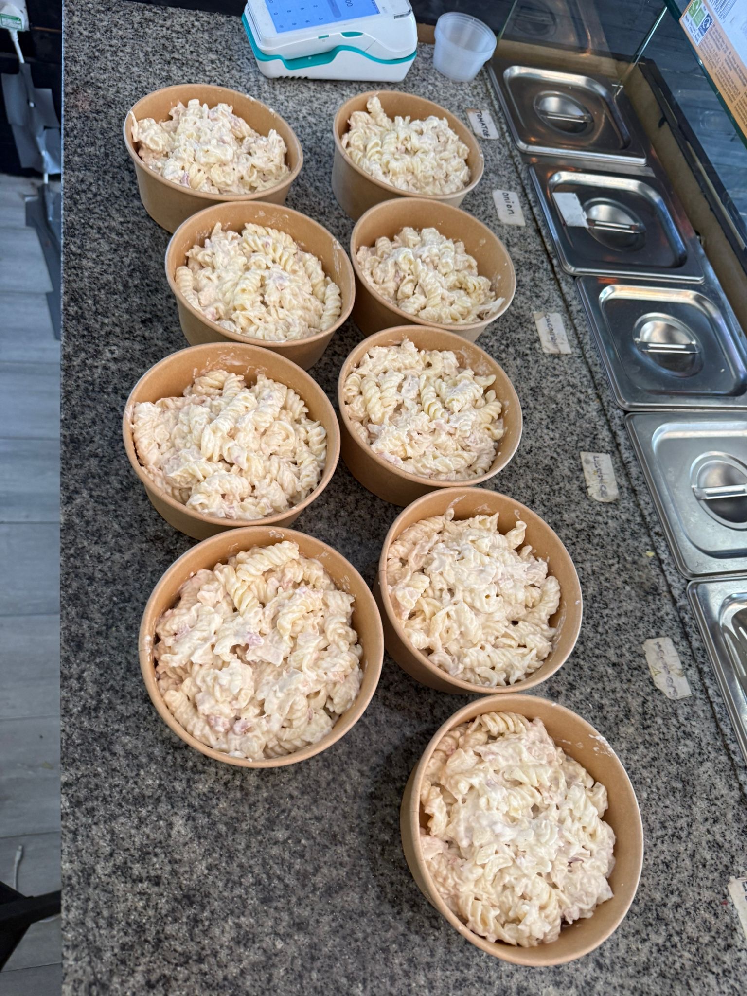 “Nine meal prep bowls filled with creamy pasta salad arranged on a granite counter in a food prep area, with ingredient trays and a payment terminal visible in the background.”