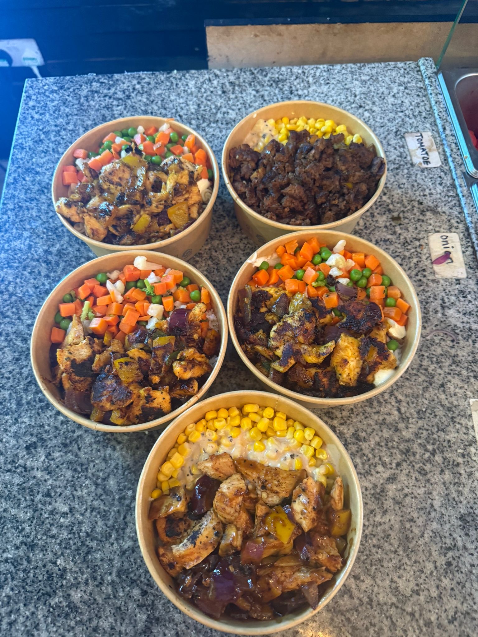 Five assorted meal prep bowls displayed on a counter. Each bowl features different combinations of grilled chicken, beef, mixed vegetables (carrots, peas, sweetcorn, green beans), and rice.
