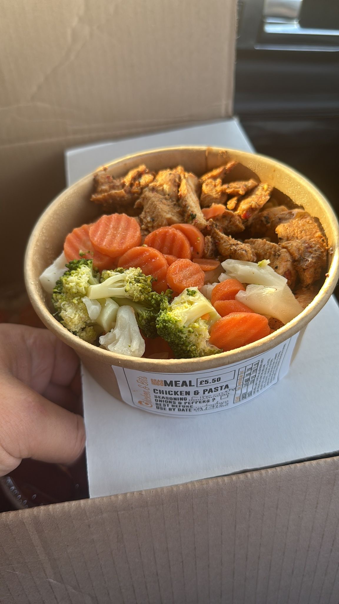 A meal prep bowl with grilled chicken, steamed broccoli, carrots, and cauliflower, placed inside a box. The outer product label reads “Chicken & Pasta” with ingredients and price information.