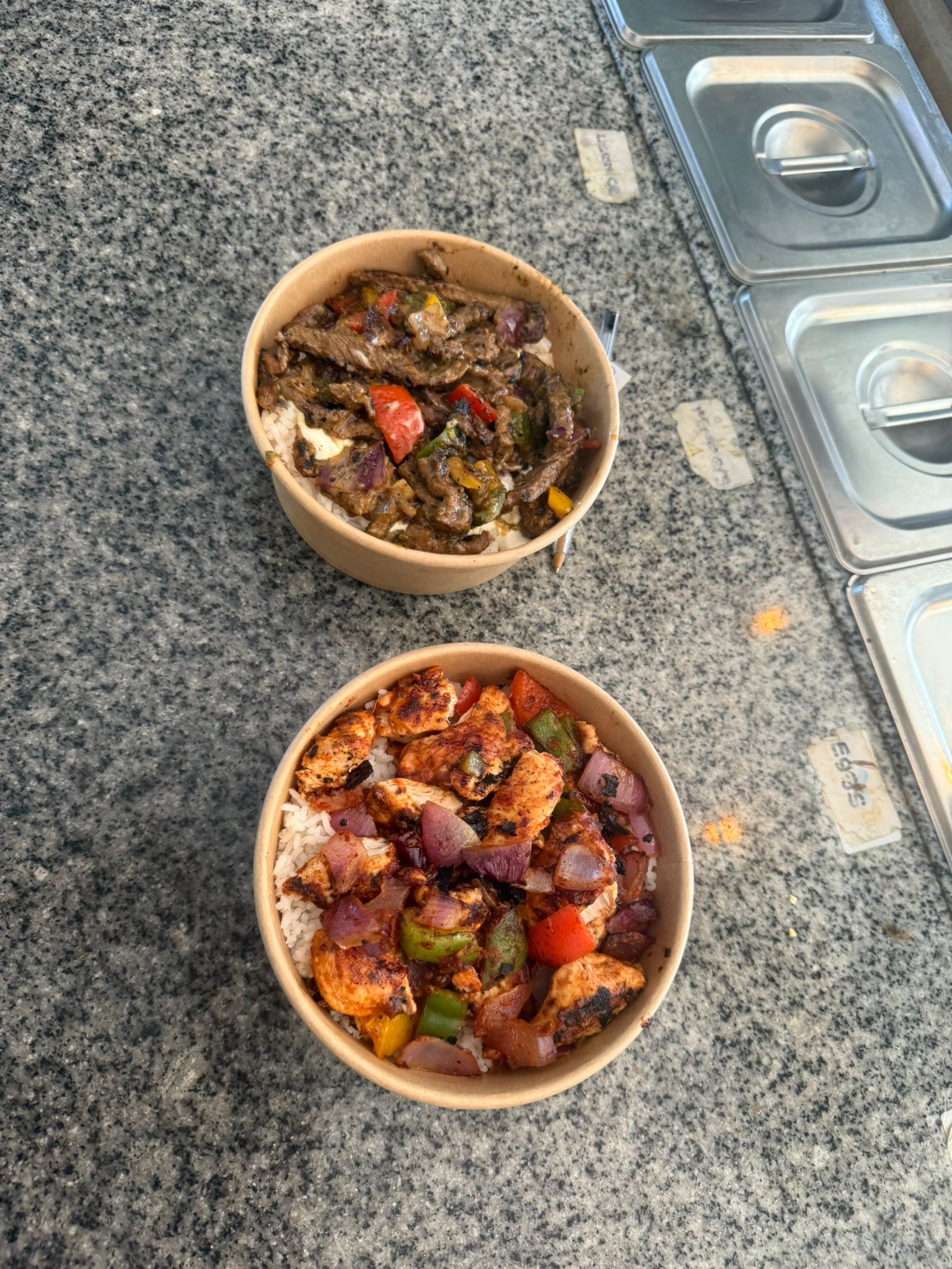 Two meal prep bowls on a granite counter. The top bowl contains sliced steak with mixed peppers over rice. The bottom bowl contains seasoned grilled chicken with mixed peppers over white rice.