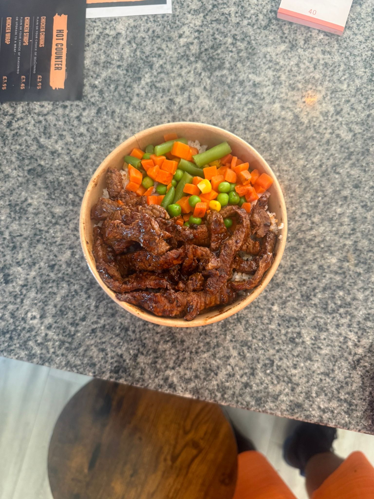 A bowl featuring strips of marinated steak with mixed vegetables including carrots, peas, green beans, and sweetcorn, served over rice on a granite counter.