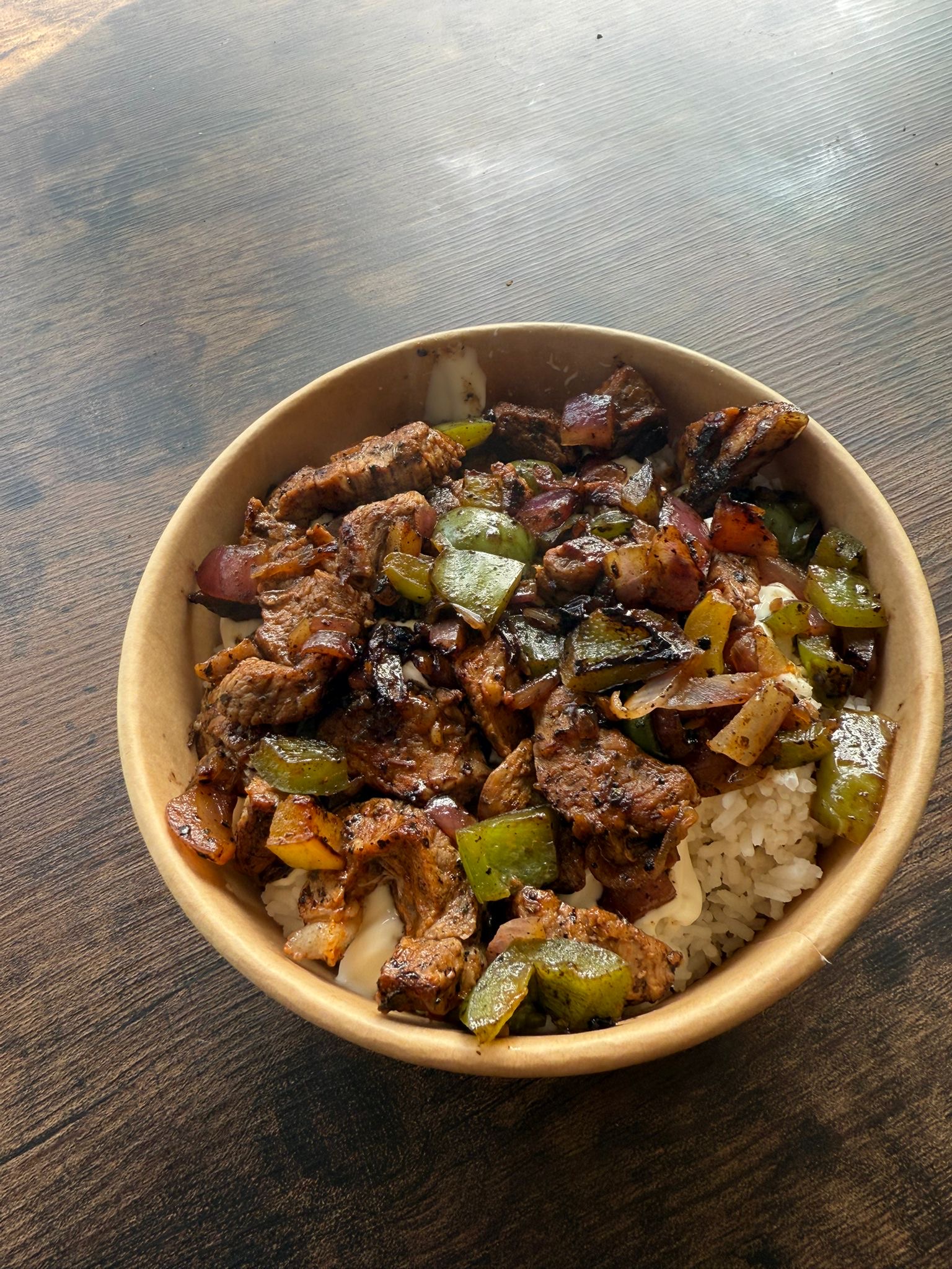 A hearty bowl of marinated grilled steak with green peppers and red onions served over white rice, presented on a wooden table.