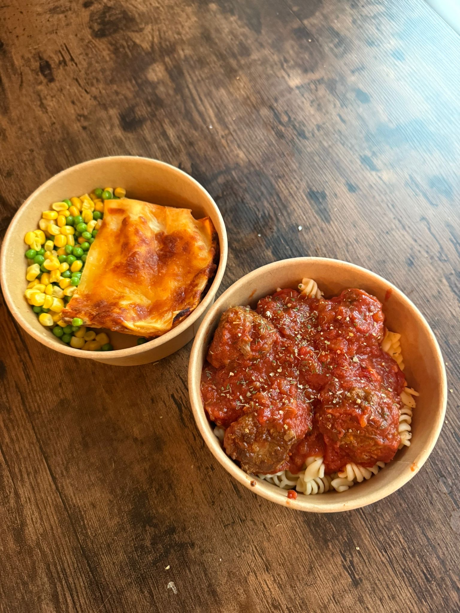 A pair of meal prep bowls on a rustic wooden table. One bowl contains pasta topped with rich tomato sauce and meatballs. The second bowl features a slice of lasagna served with peas and sweetcorn.