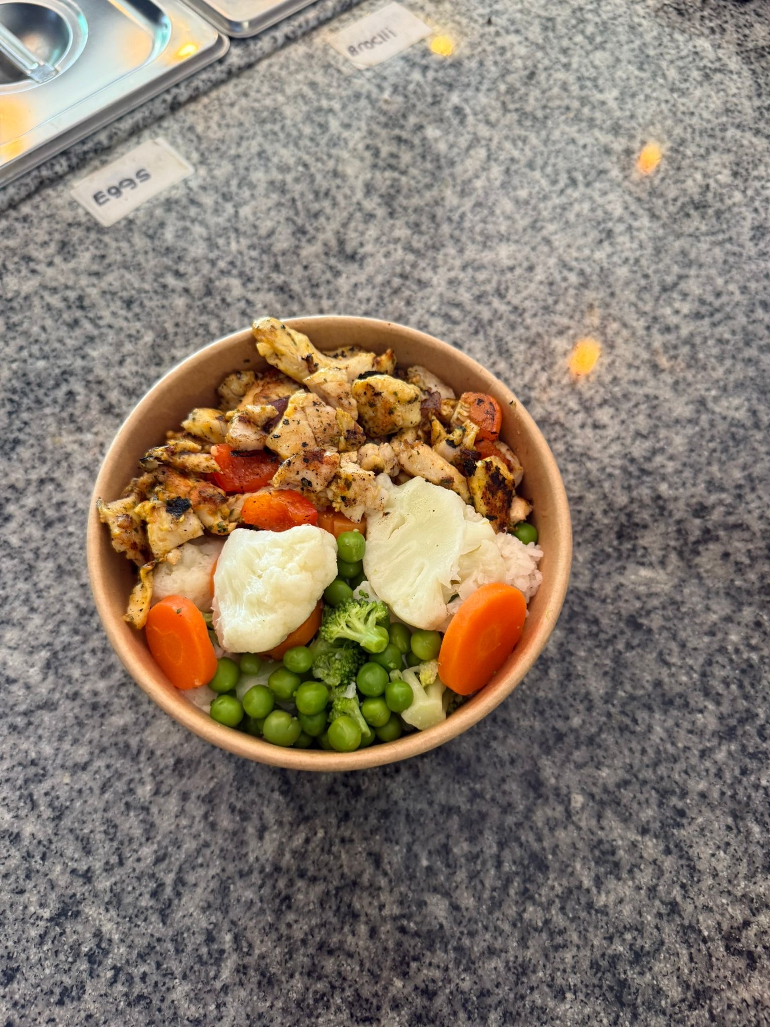 A bowl filled with lean grilled chicken pieces, steamed carrots, peas, cauliflower, and broccoli, placed on a prep counter.