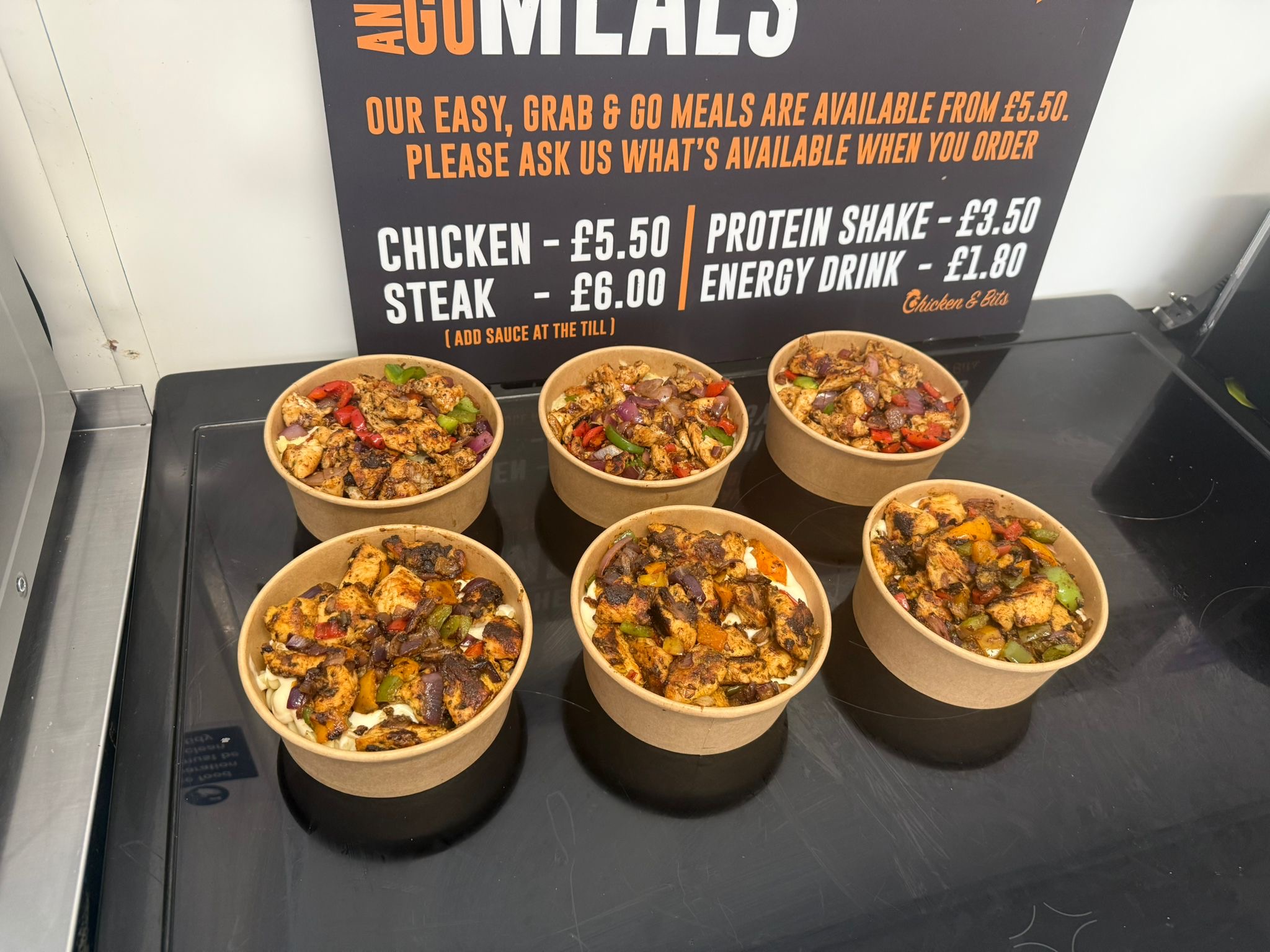 Six freshly prepared meal prep bowls arranged in front of a “Grab & Go Meals” sign. Each bowl contains grilled chicken or steak with colourful diced vegetables and rice, showcasing the ready-to-go menu options.