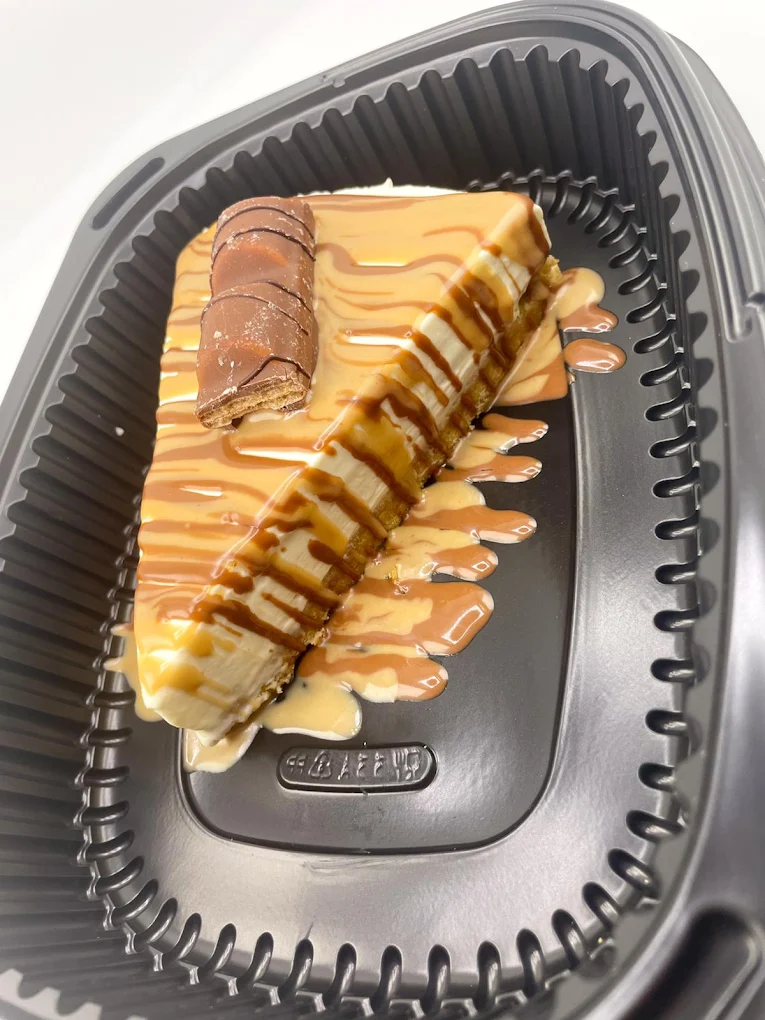 Creamy cheesecake slice topped with caramel and white chocolate sauce, served in a black takeaway tray.