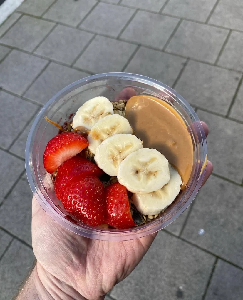 Fresh fruit bowl with sliced banana, strawberries, crunchy granola and a drizzle of peanut butter, presented in a clear takeaway container.