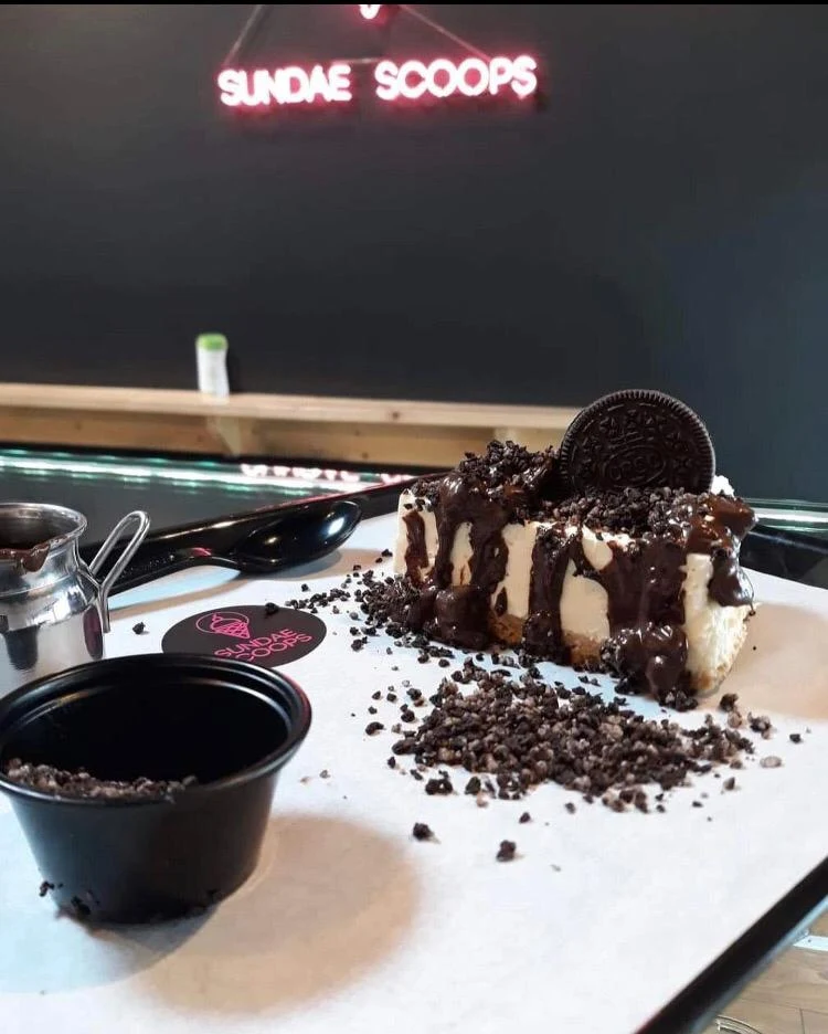 Cheesecake slice topped with crushed Oreo cookies, chocolate sauce and a whole Oreo biscuit, served on a branded tray.