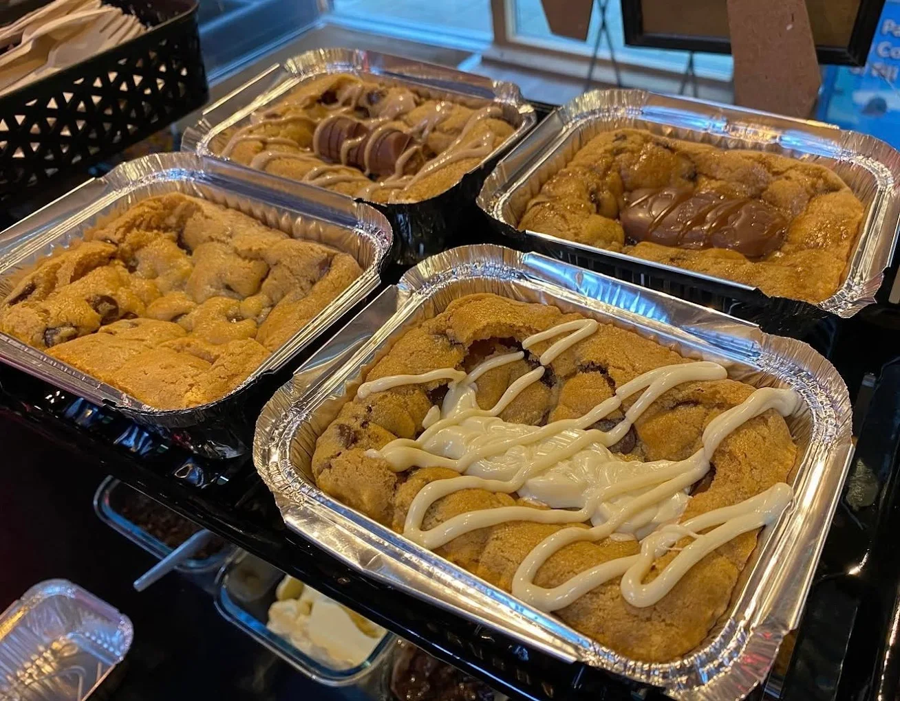 Freshly baked cookie dough served in foil trays, topped with chocolate bars and white chocolate drizzle, displayed ready to serve.