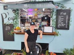 Owner, Lisa, standing in front of the food truck hatch. Coffee menu on display with baked goods and condiments. Daily special board also in view.