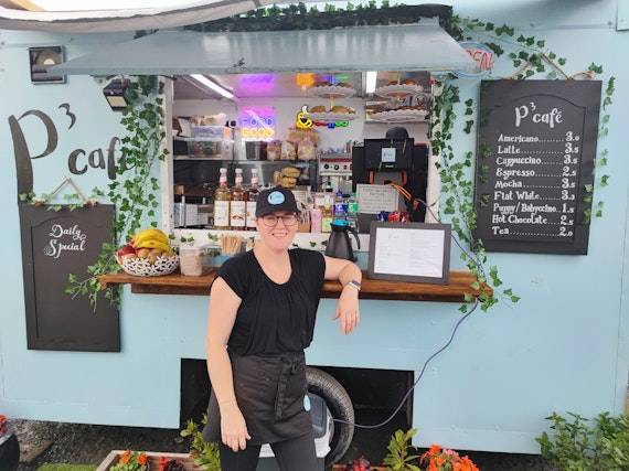 Owner, Lisa, standing in front of the food truck hatch. Coffee menu on display with baked goods and condiments. Daily special board also in view.